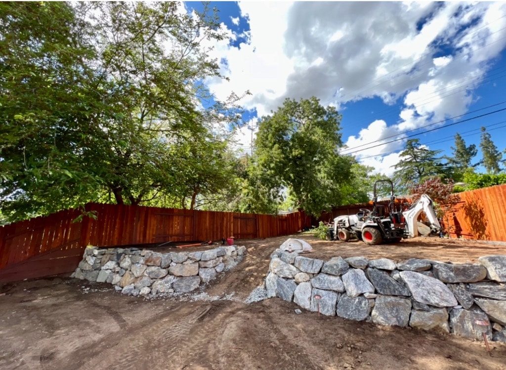Boulder retaining walls under construction with Bobcat excavator in backyard